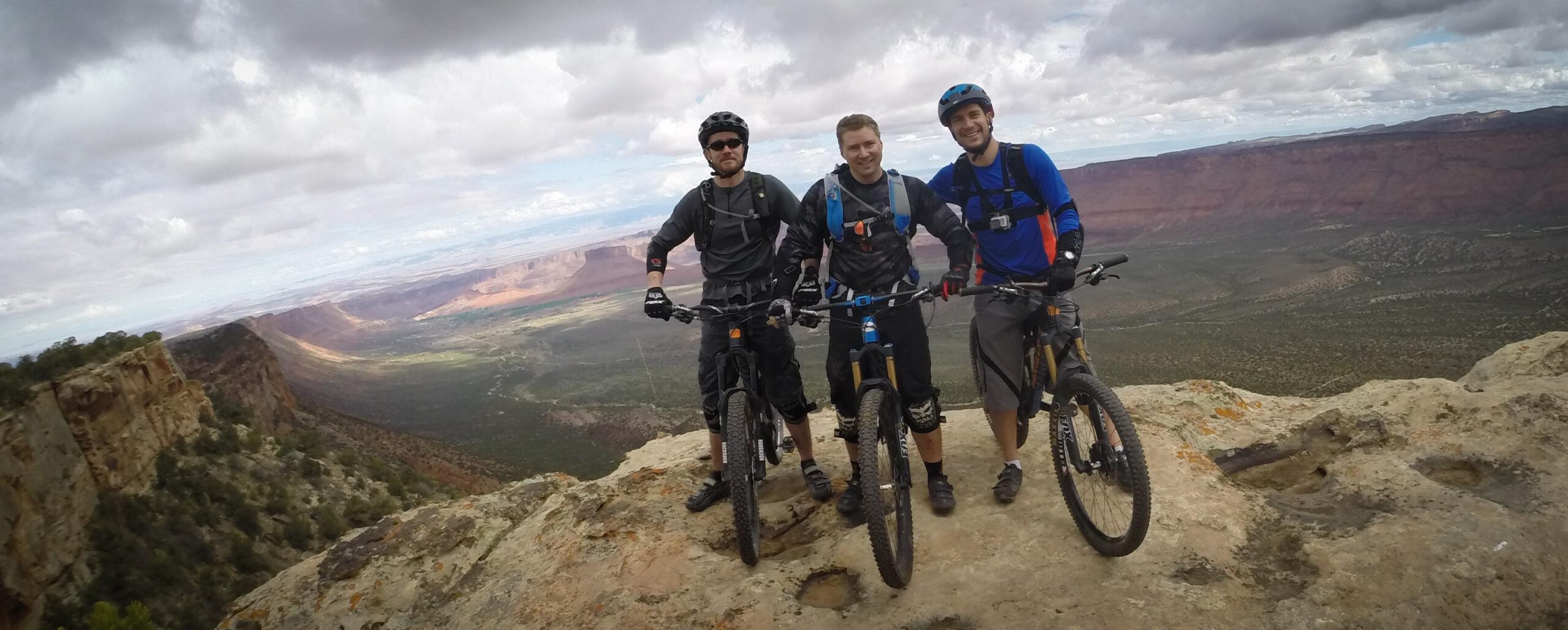 Three mountain bikers pose on a rocky ledge with a vast scenic landscape in the background. The sky is overcast, adding a dramatic effect to the view of rolling hills and distant cliffs. Each rider is wearing protective gear and standing next to their bikes, smiling towards the camera. Porcupine Rim mountain bike trail.