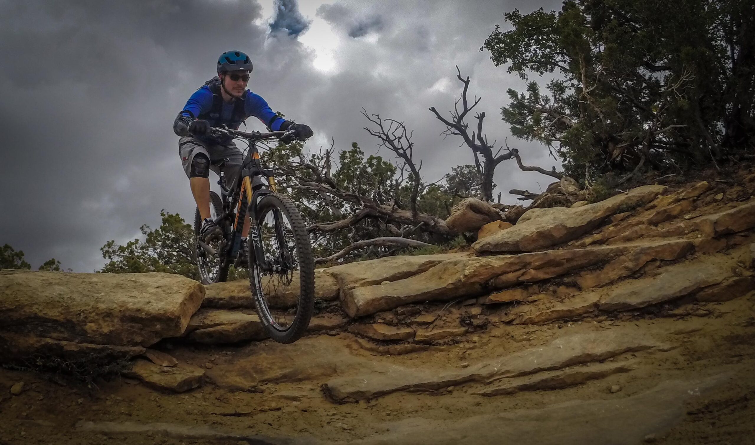 A mountain biker navigating a rocky trail, wearing a blue long-sleeve shirt and a helmet, with overcast skies in the background. Trees and rocky terrain surround the scene. Porcupine Rim mountain bike trail.
