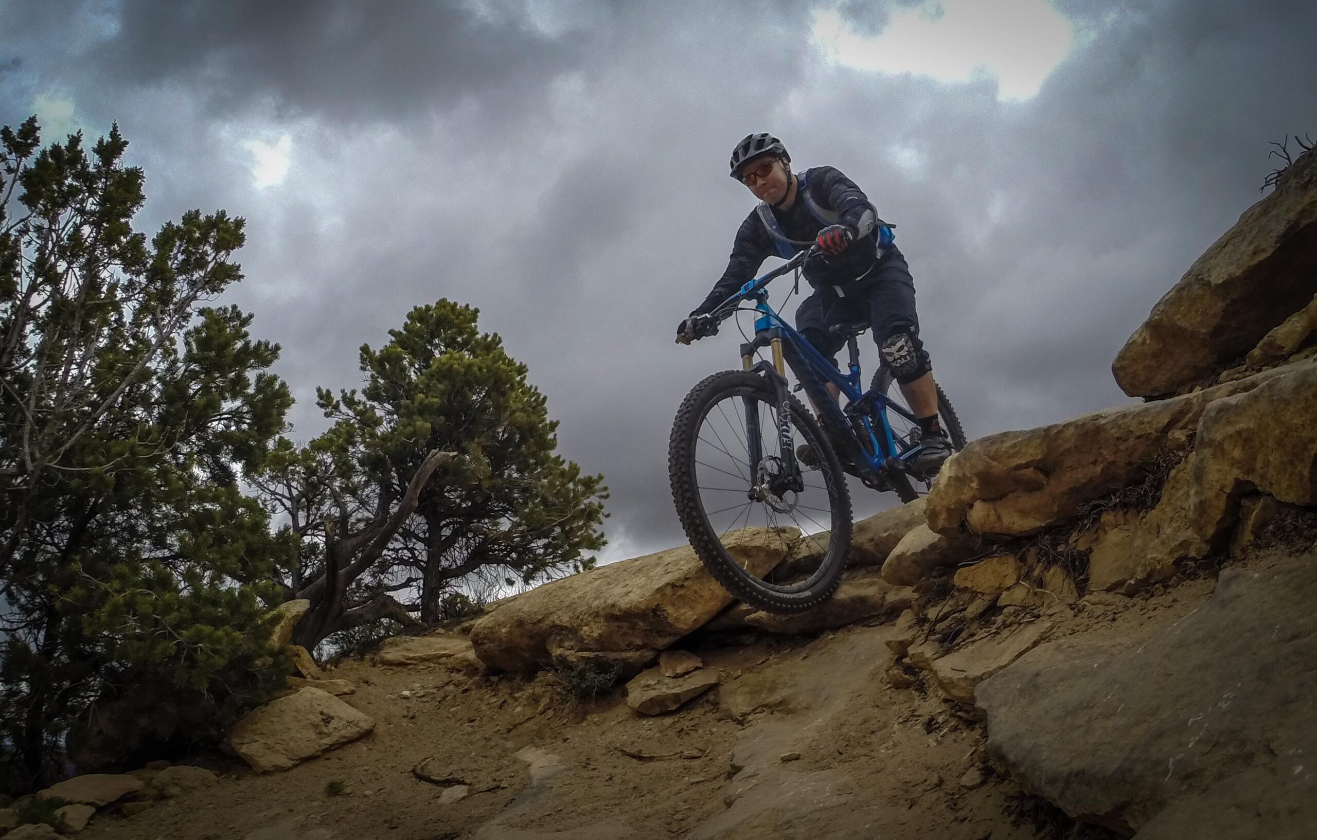 A mountain biker navigating a rocky trail, leaning forward on a blue mountain bike, with cloudy skies and trees in the background. Porcupine Rim mountain bike trail.