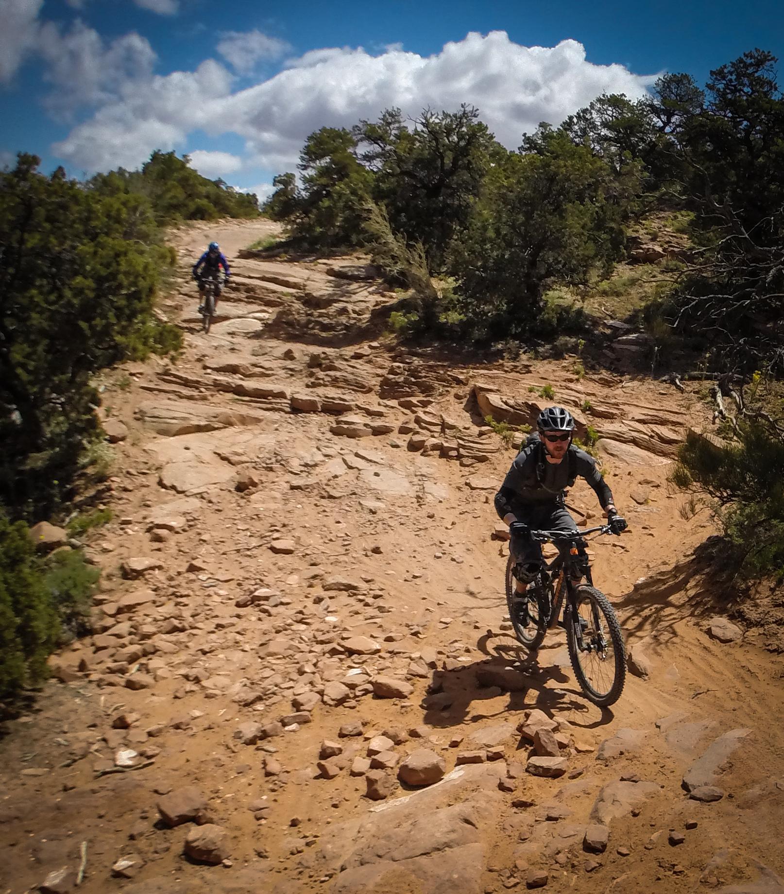 Two mountain bikers navigate a rocky trail, surrounded by greenery and a blue sky with clouds. One biker is descending a steep section while the other is further up the trail. The terrain is uneven, with visible rocks and dirt. Porcupine Rim mountain bike trail.
