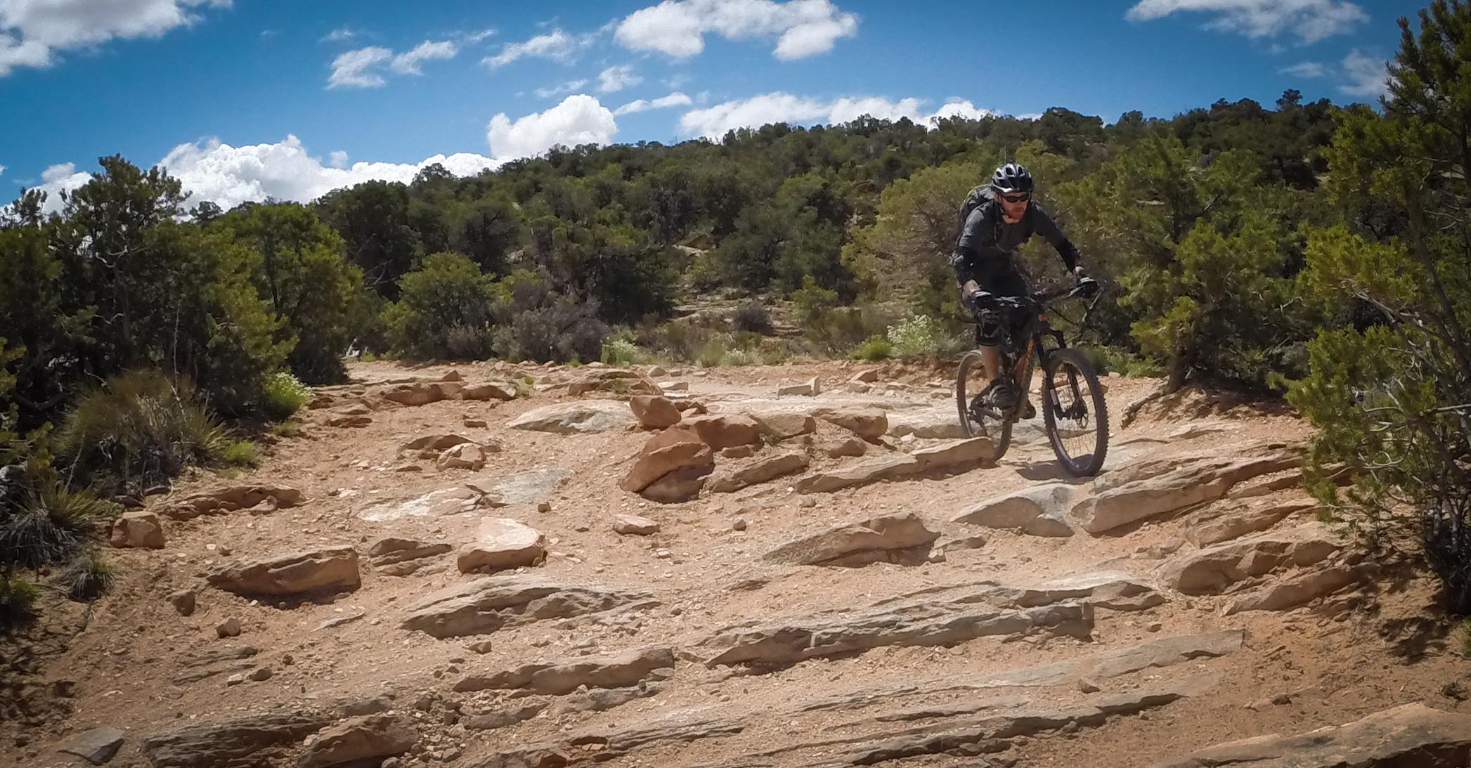 A mountain biker navigating a rocky trail surrounded by green vegetation and blue skies with scattered clouds. Porcupine Rim mountain bike trail.