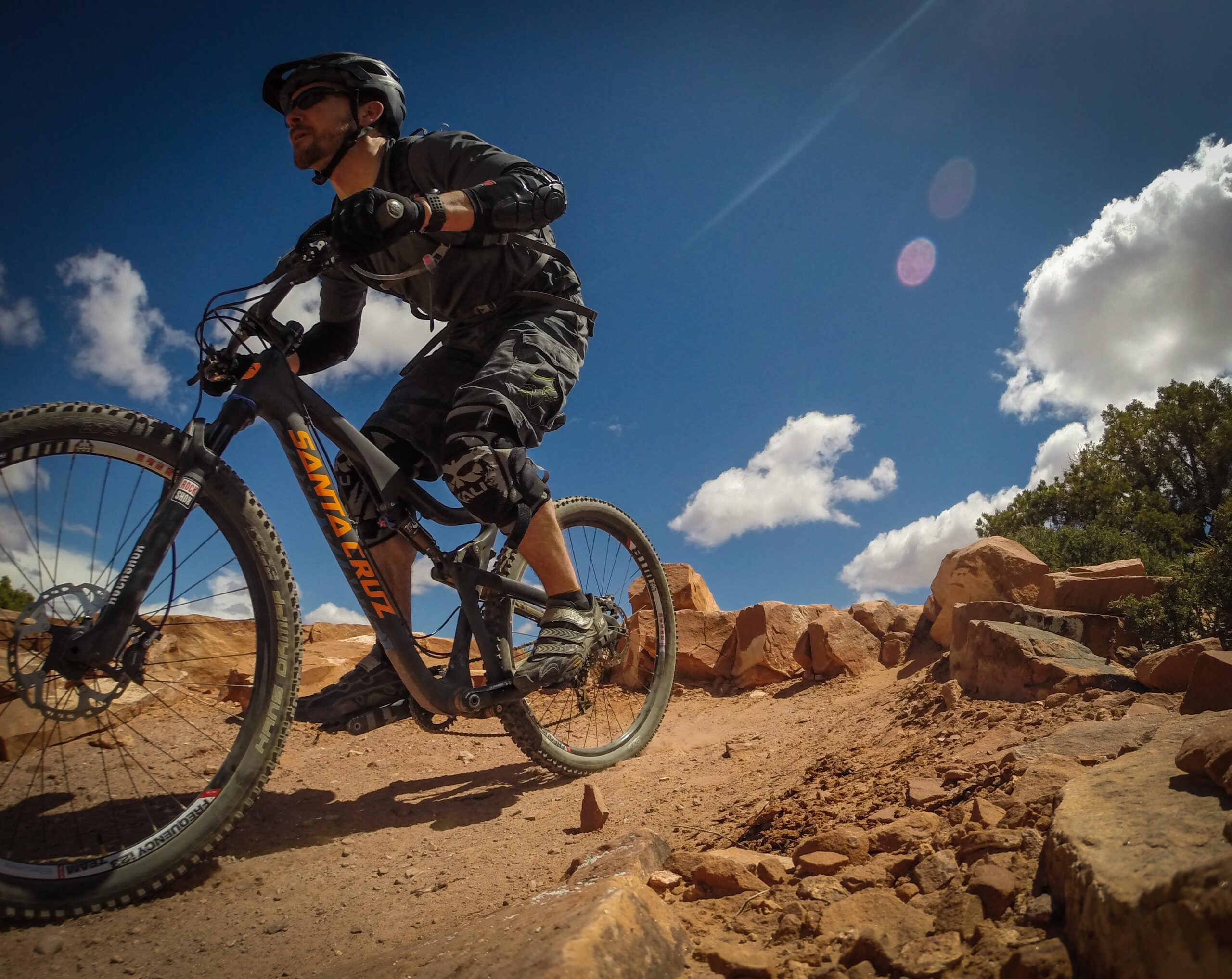 A mountain biker riding on rocky terrain under a bright blue sky with scattered clouds. The cyclist is wearing a helmet and protective gear, showcasing a dynamic riding posture. Porcupine Rim mountain bike trail.