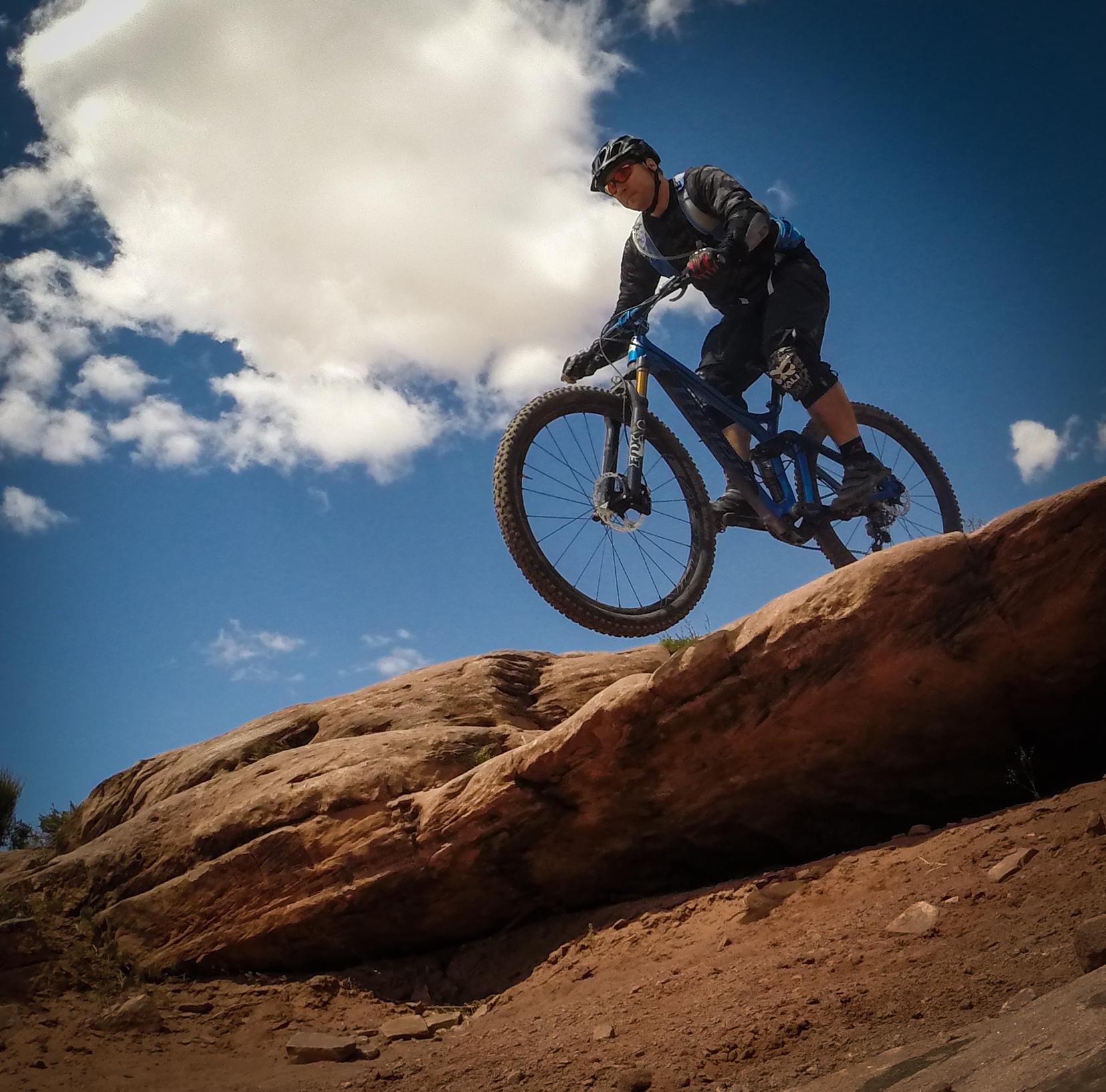 A mountain biker jumping off a rock formation against a blue sky with clouds. The biker is wearing a helmet and protective gear, and is captured mid-air while riding a blue mountain bike. Porcupine Rim mountain bike trail.