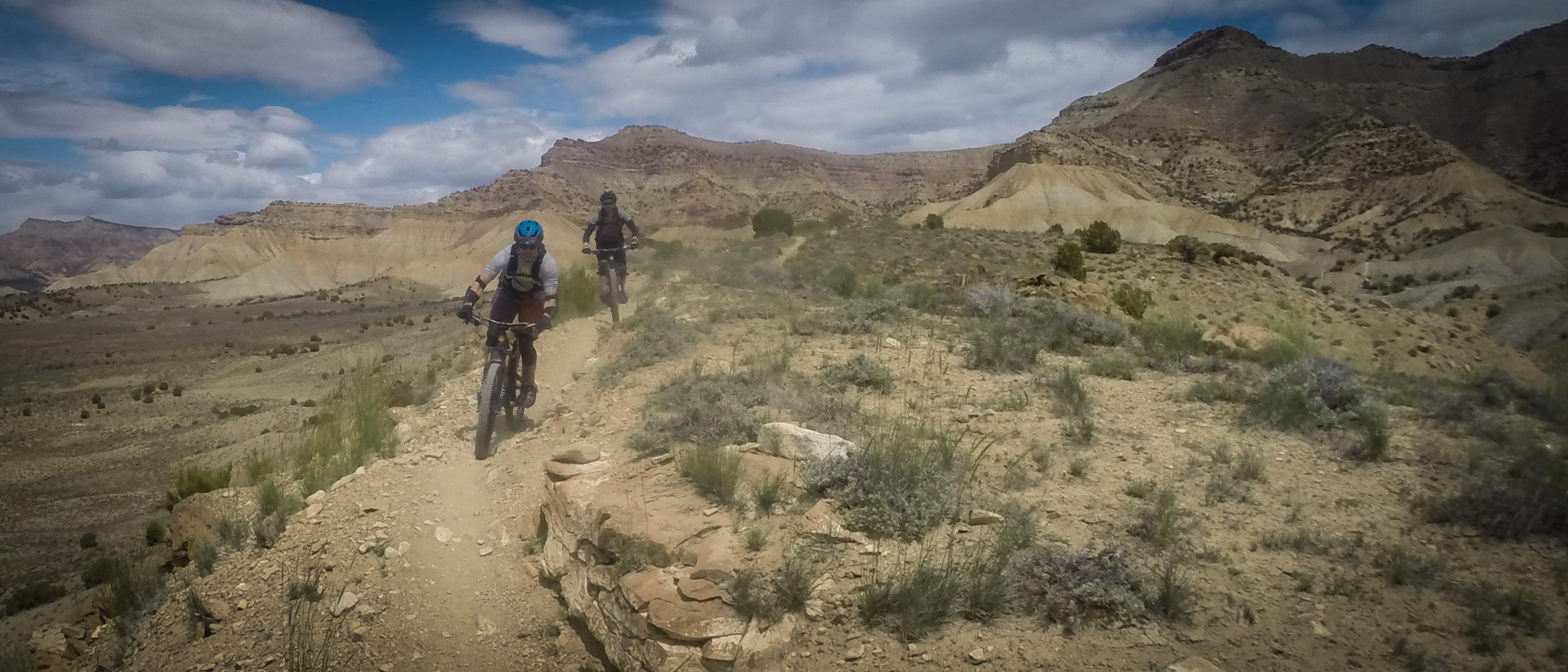 Two mountain bikers riding along a narrow dirt trail in a rugged, rocky landscape with hills and low vegetation under a partly cloudy sky. Zippety Do Dah mountain bike trail.