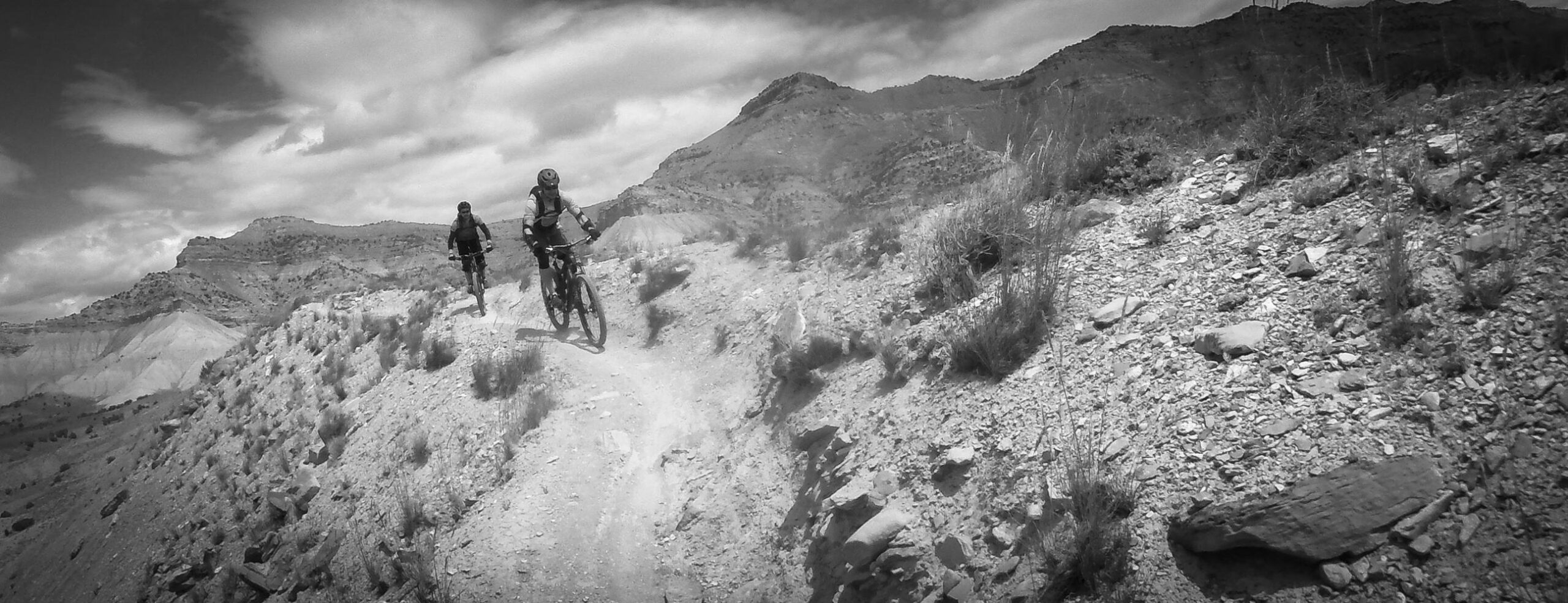 Two mountain bikers ride along a narrow dirt trail on a rocky terrain, surrounded by rugged hills and cloudy skies. The scene is captured in black and white, emphasizing the natural landscape and the riders’ dynamic movement. Zippety Do Dah mountain bike trail.