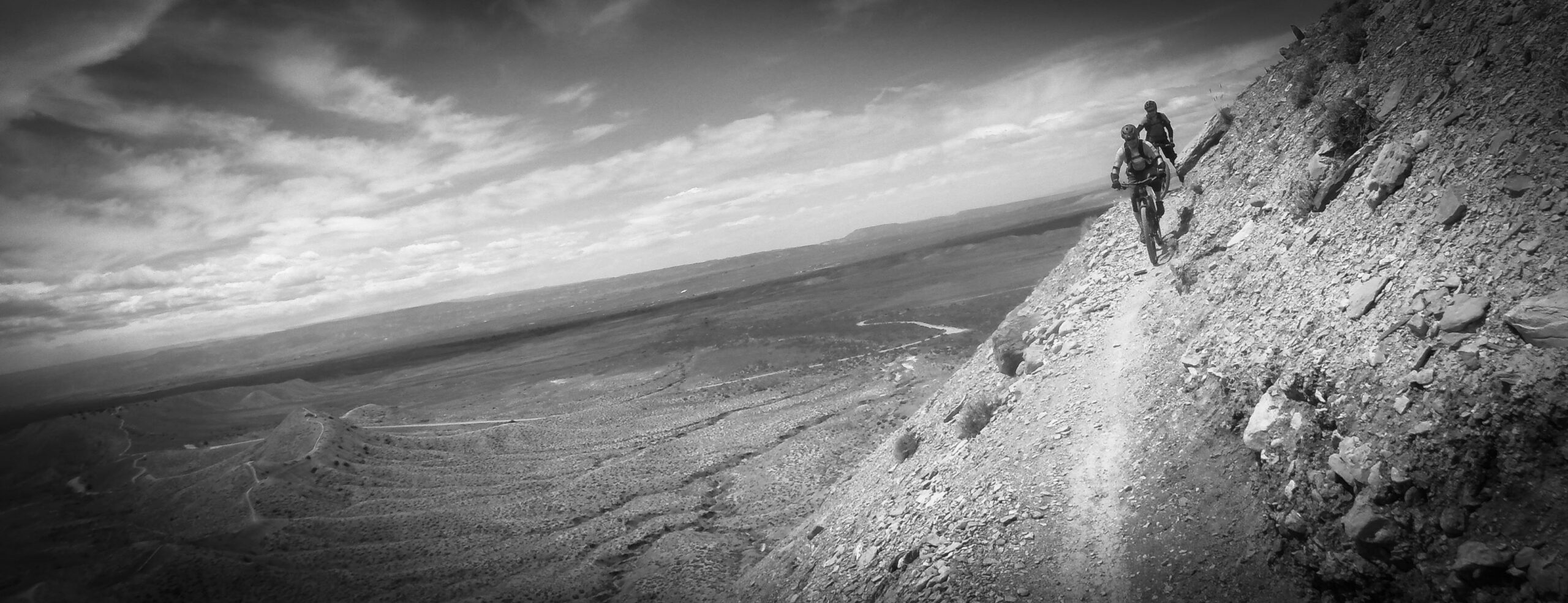 A mountain biker riding along a narrow, rocky trail on a steep terrain, with a panoramic view of a valley and distant hills under a cloudy sky. The image is in black and white, emphasizing the rugged landscape and the challenging biking conditions. Zippety Do Dah mountain bike trail.