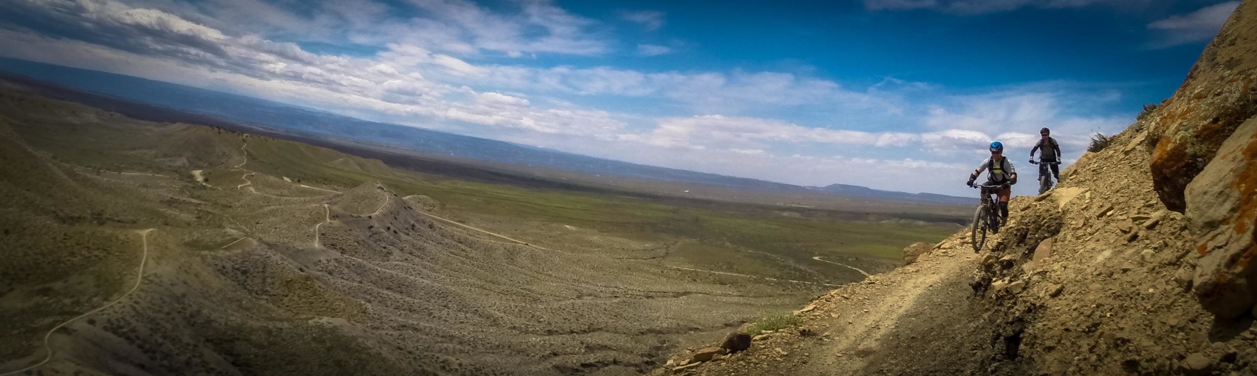 Two mountain bikers navigate a rocky trail along a steep cliff, with expansive green valleys and a cloudy blue sky in the background. The landscape features rolling hills and distant mountains, showcasing an adventurous outdoor setting. Zippety Do Dah mountain bike trail.