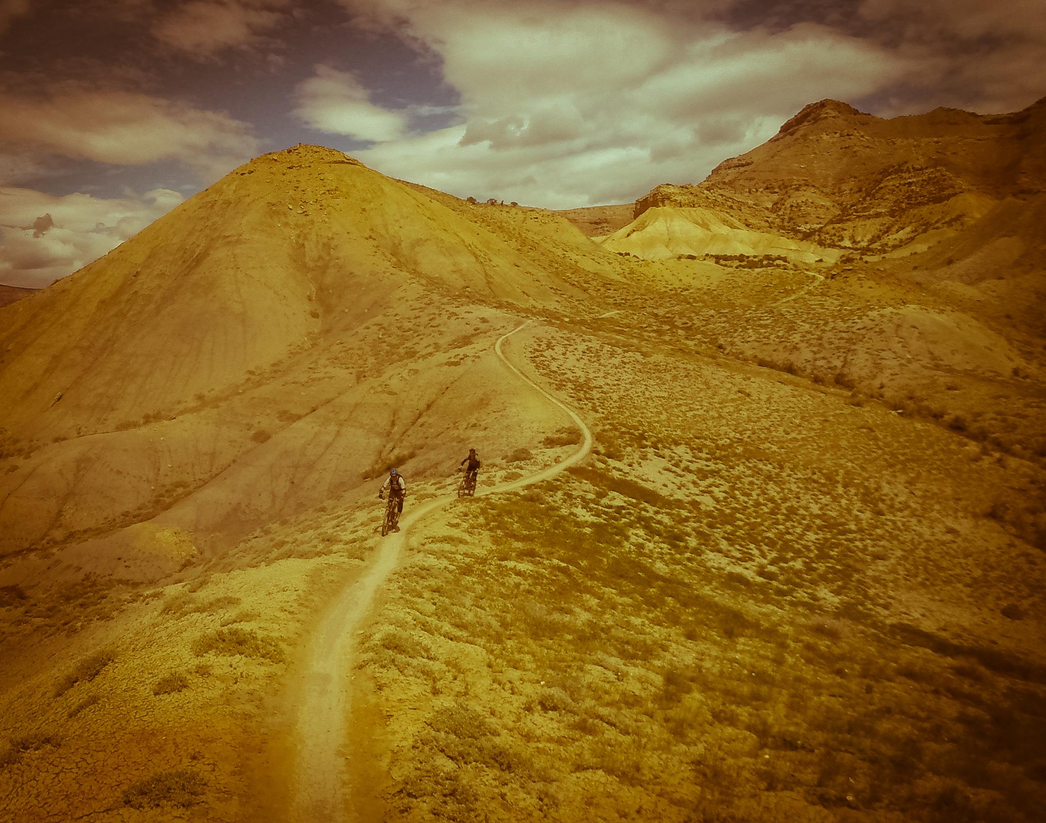 Two mountain bikers riding on a dirt trail winding through arid, hilly terrain under a cloudy sky. The landscape features shades of brown and green, with textured hills and sparse vegetation. Zippety Do Dah mountain bike trail.