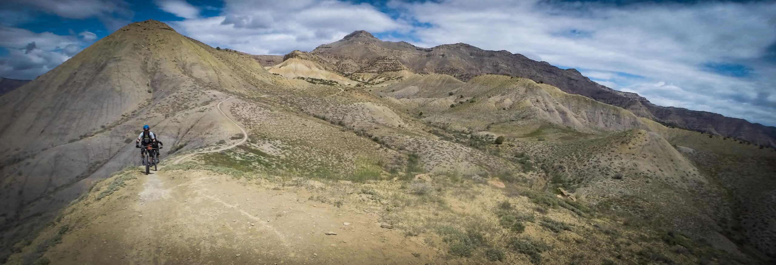 A cyclist navigating a rocky mountain trail surrounded by rugged hills and a partly cloudy sky. The landscape features varying shades of brown and green with visible dirt paths winding through the terrain. Zippety Do Dah mountain bike trail.