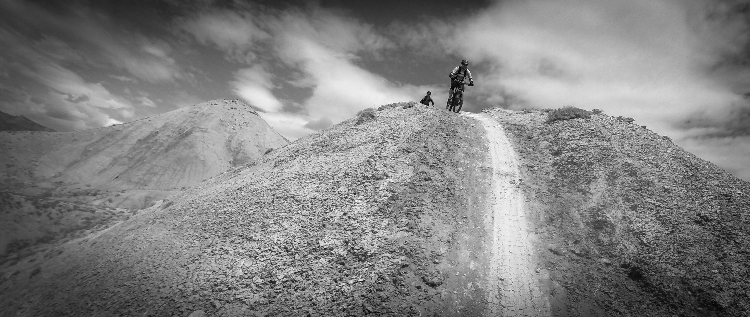 A black and white photograph of two mountain bikers on a rugged hillside, with one rider descending a narrow dirt path and the other standing nearby. The landscape features steep, rocky terrain and distant mountains under a partly cloudy sky. Zippety Do Dah mountain bike trail.