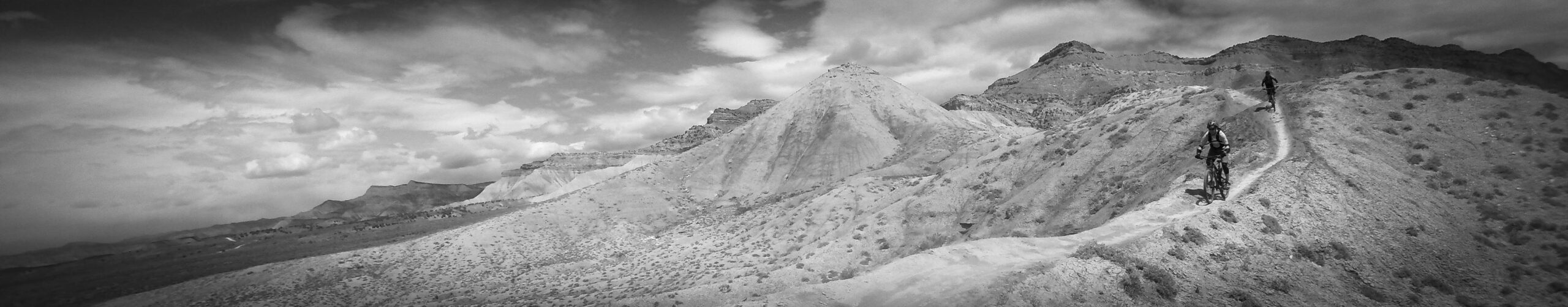 A black and white panoramic image of two mountain bikers riding along a winding dirt trail through a rugged, arid landscape. The background features rocky hills and cloud-patterned skies, emphasizing the vastness of the terrain. Zippety Do Dah mountain bike trail.