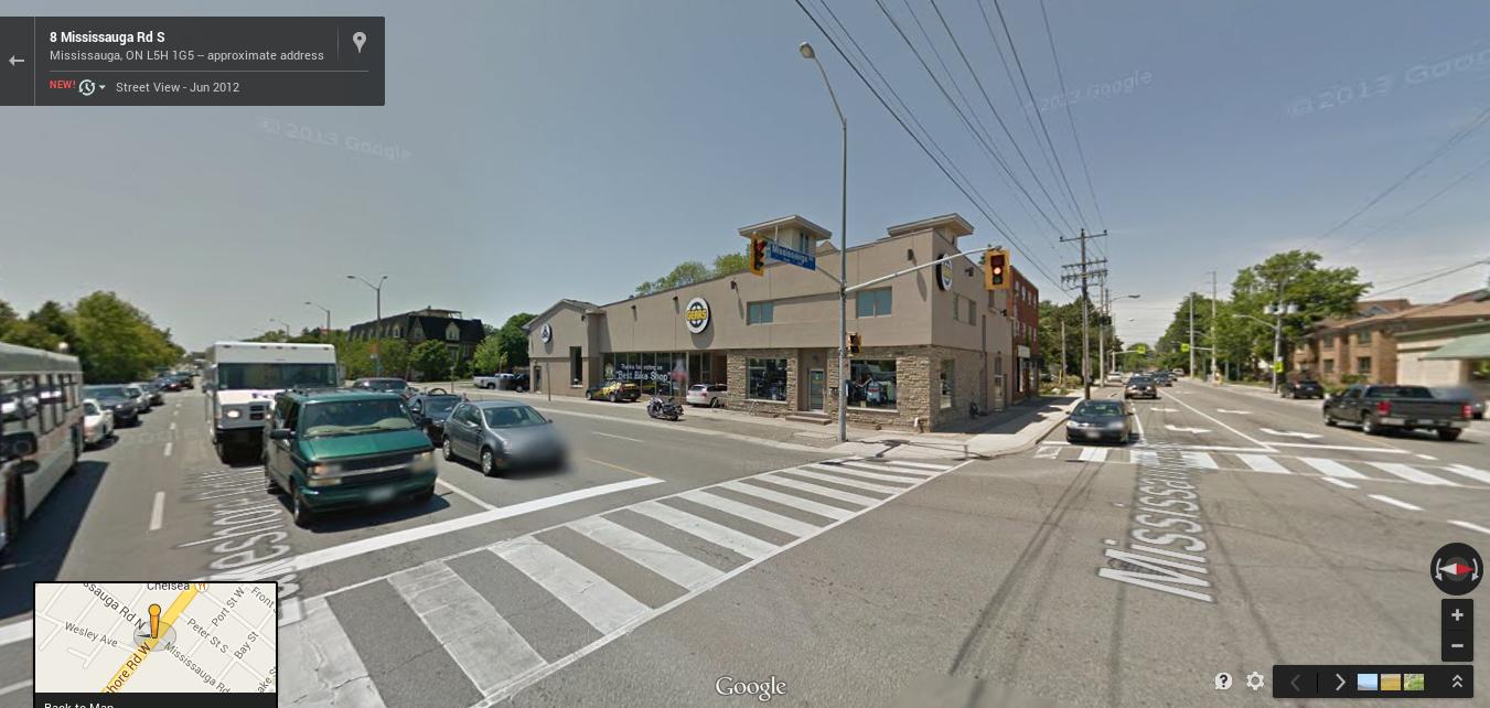 A street view image showing an intersection on Mississauga Road South. The foreground features a crosswalk with several vehicles, including buses and cars. On the right side, there is a commercial building with large windows displaying a business sign. Street signs and traffic lights are visible, along with trees lining the streets. The image captures a sunny day in an urban setting.