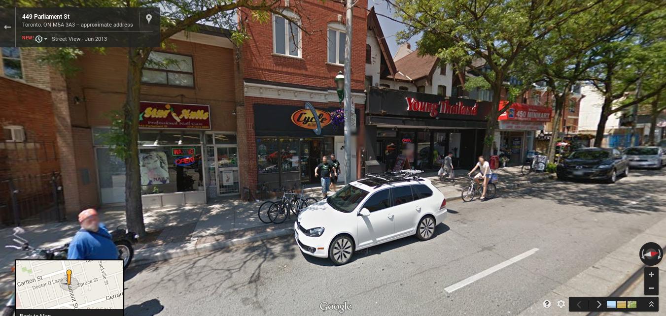 A busy street scene on Parliament Street in Toronto, featuring several storefronts including a nail salon and a bike shop. A white car is parked along the road, and pedestrians, including a person on a bicycle, are seen walking by. Lush trees provide shade along the sidewalk. A map inset is visible in the corner.