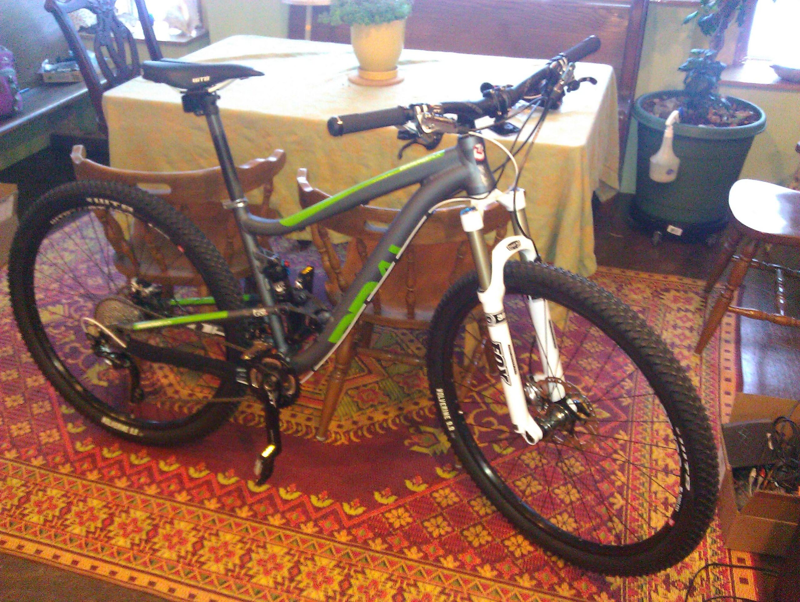 Diamondback Sortie 3 29er: A mountain bike with a gray and green frame is positioned in a cozy room, surrounded by wooden furniture and a colorful patterned rug. The bike features a white front fork, thick tires, and a seat that appears adjustable. In the background, there is a dining table covered with a cloth and potted plants.