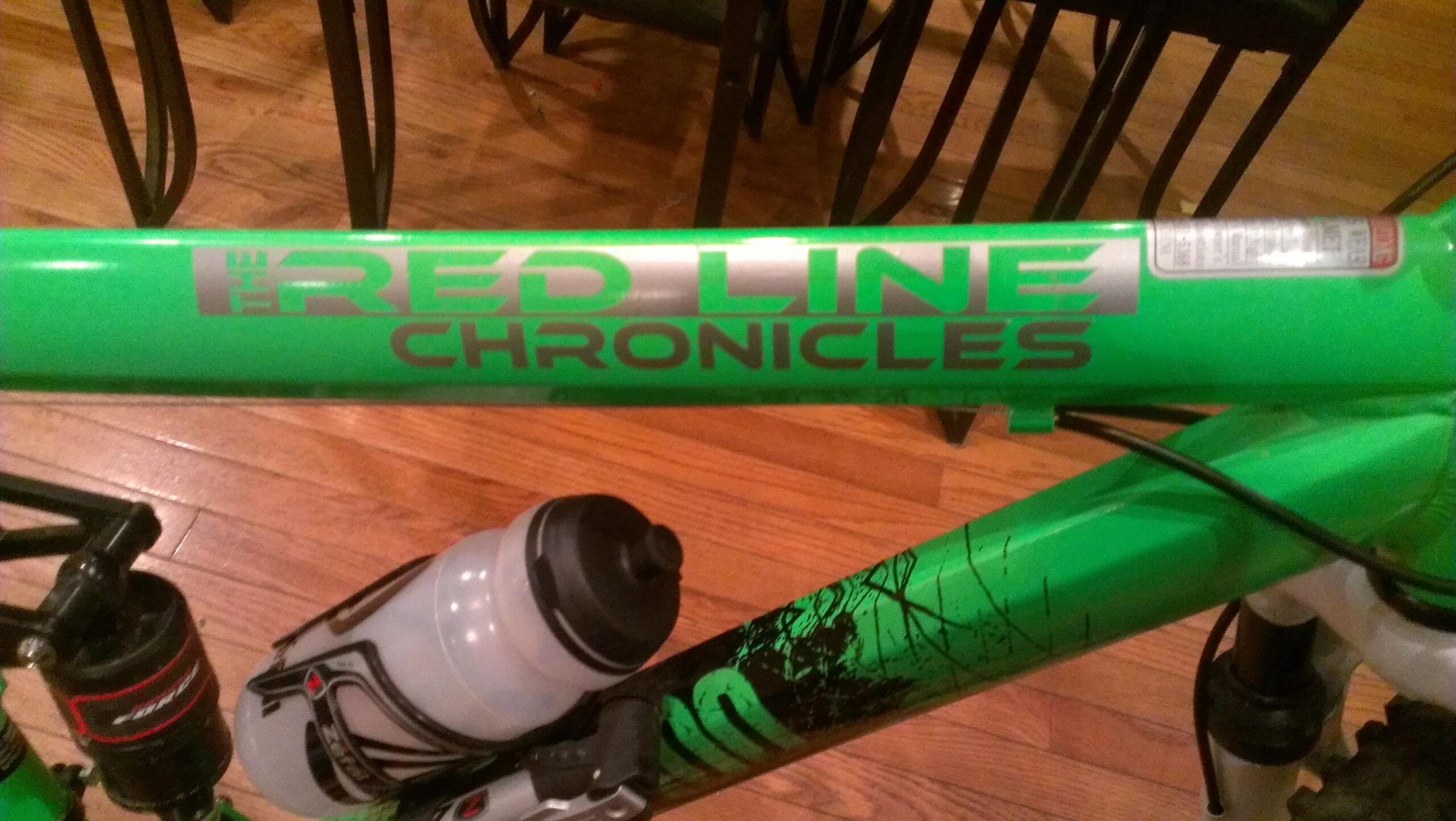 Close-up view of a vibrant green bicycle frame featuring the text "The Red Line Chronicles." A water bottle is attached to the frame, with wooden flooring in the background.