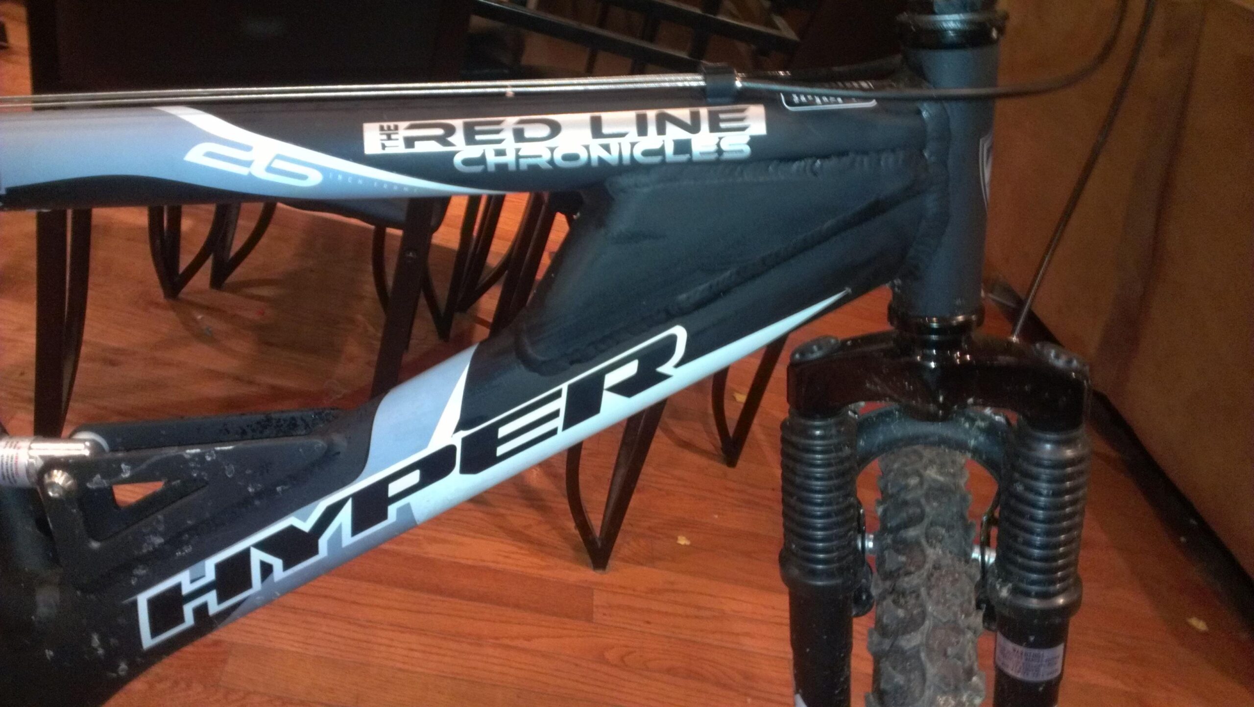Mountain bike with a prominently displayed "Hyper" logo and the phrase "The Red Line Chronicles" on its frame, positioned on a wooden floor. The bike features a front suspension with some mud on the tires, indicating recent use. In the background, chair legs are visible, suggesting an indoor setting.