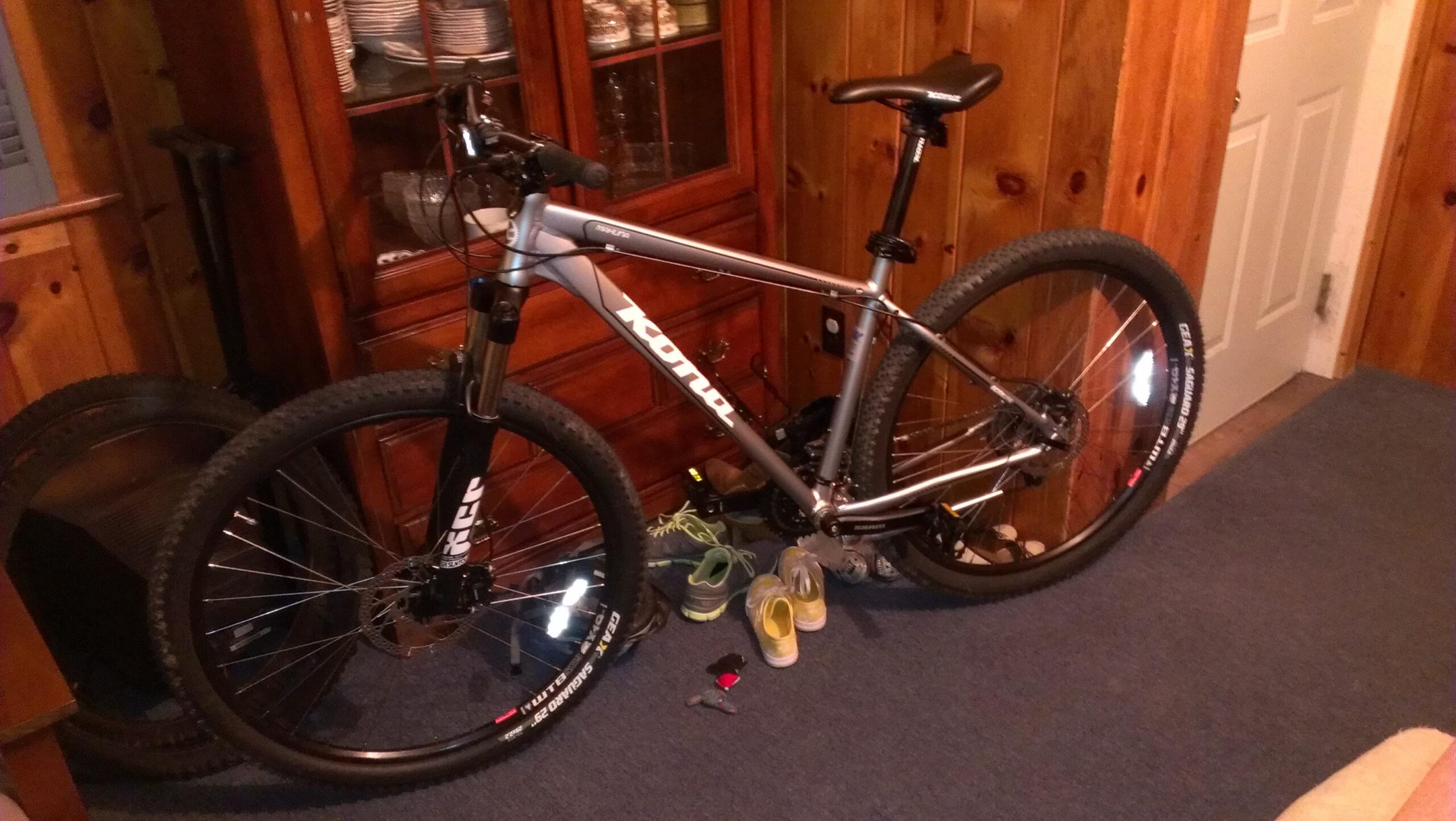 Kona Mahuna: A silver mountain bike is propped against a wooden cabinet in a room with wooden paneling. In the foreground, there are several pairs of shoes and a set of keys near the bike. The background includes a partially open door and a carpeted floor.