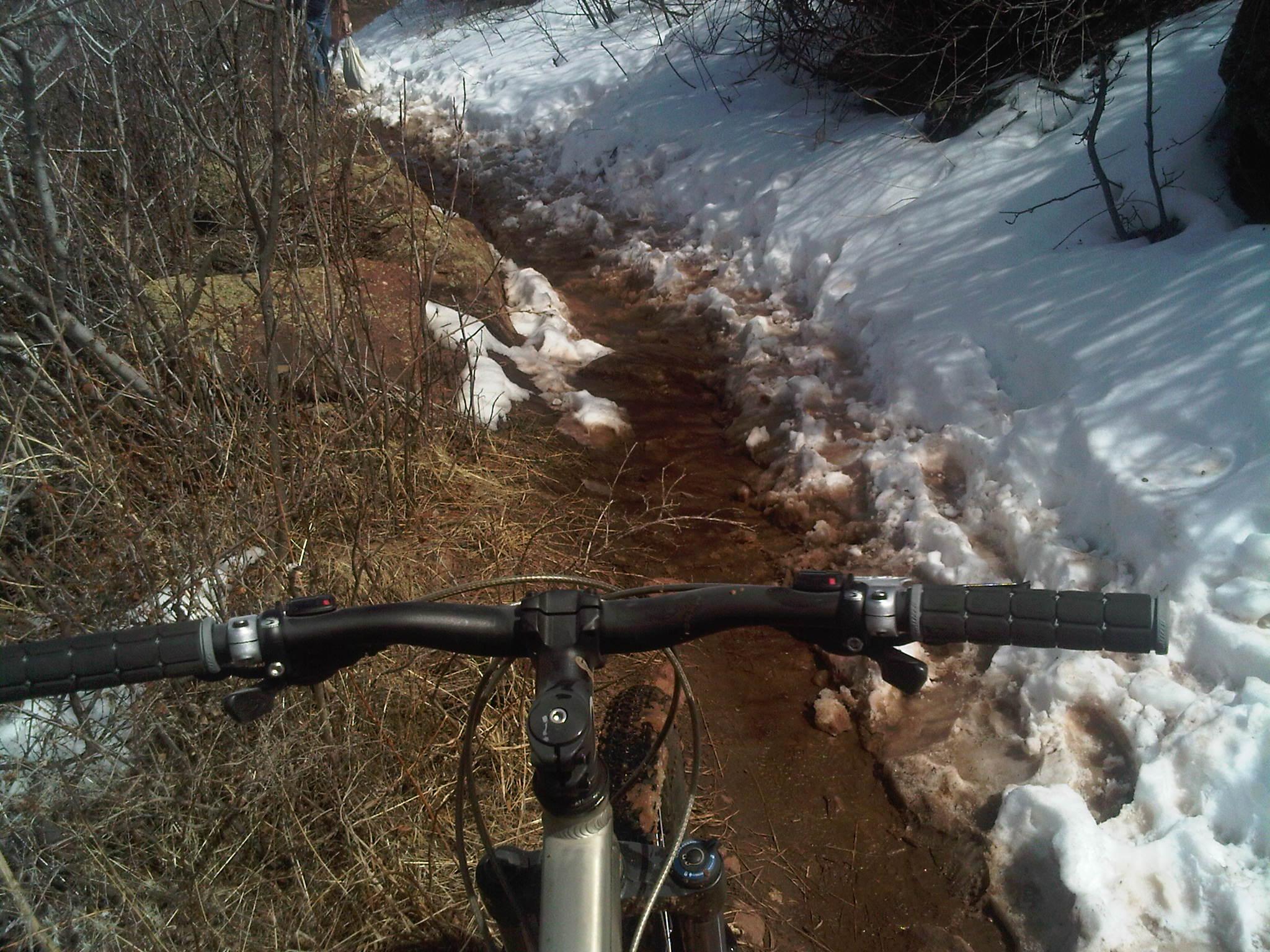 Specialized Rockhopper Comp Disc: A mountain bike's handlebars are in focus, with a dirt trail visible ahead, bordered by patches of snow and scattered grass. In the background, a person can be seen walking along the trail.