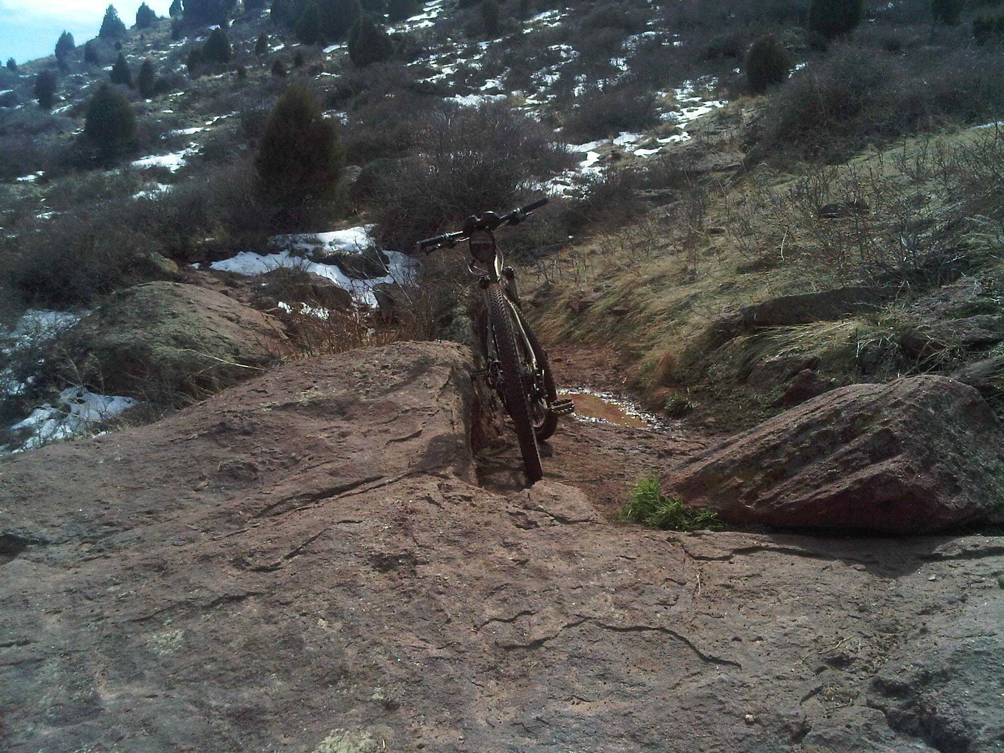 Specialized Rockhopper Comp Disc: A mountain bike leaning against a large rock on a rugged, grassy trail with sparse vegetation, scattered snow patches, and a backdrop of hilly terrain under a blue sky.