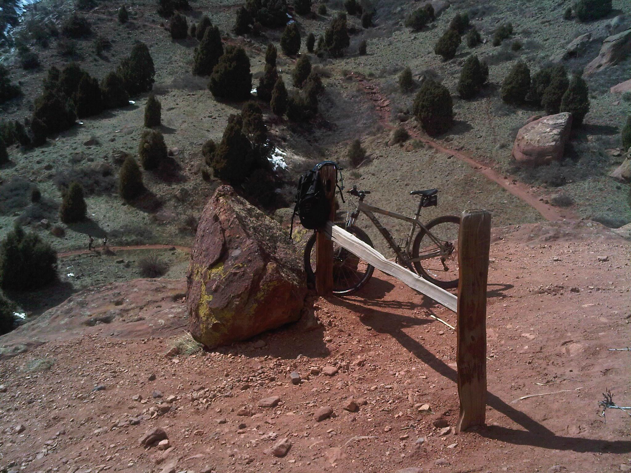 Specialized Rockhopper Comp Disc: A mountain bike leans against a wooden post on a dirt trail, overlooking a scenic landscape of shrubs and trees. A large rock is nearby, and the terrain features a mix of red soil and greenery. In the background, additional trails can be seen winding through the landscape.