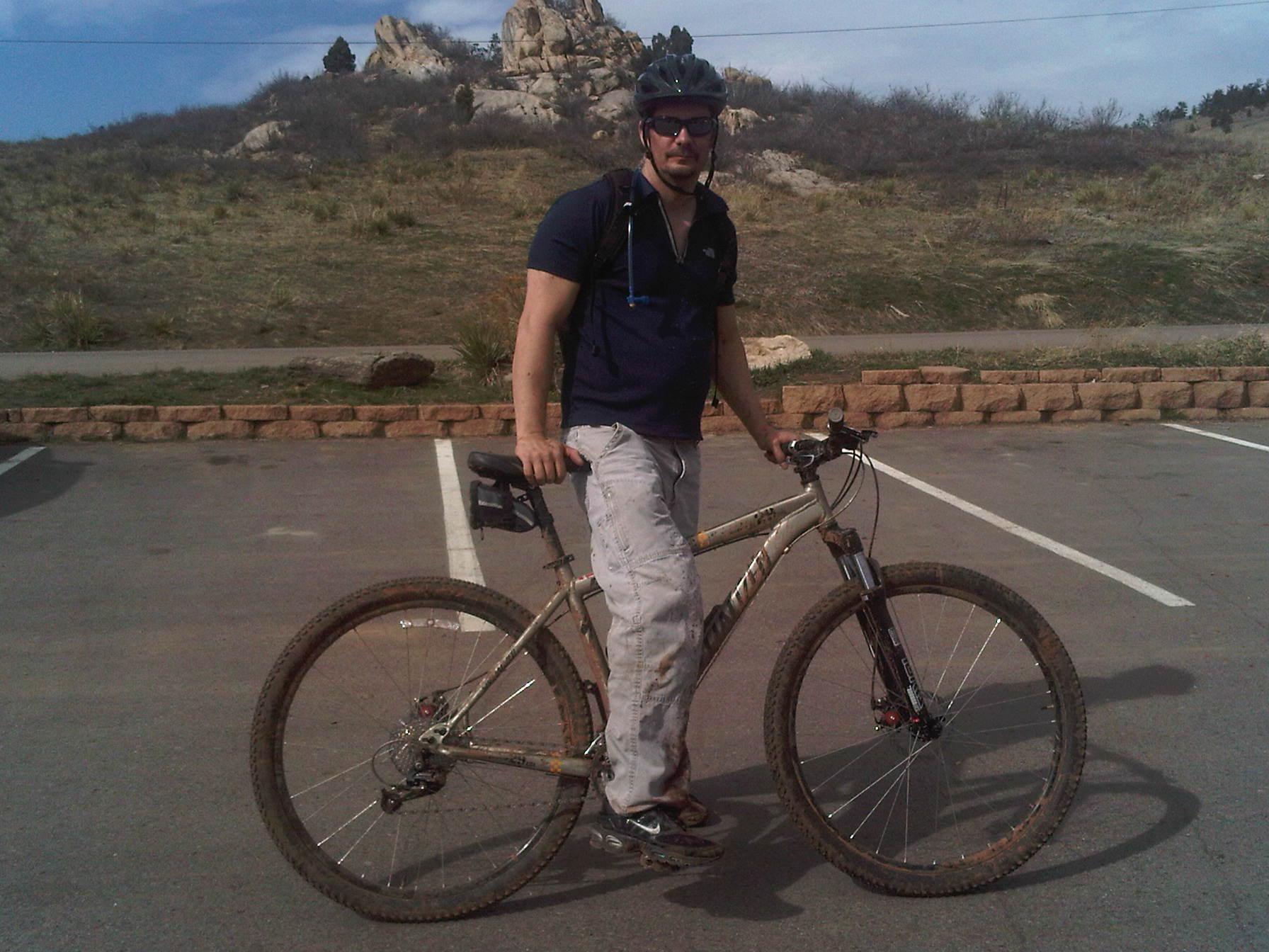 Specialized Rockhopper Comp Disc: A mountain biker stands next to a muddy bicycle in a parking lot, with rocky hills and sparse vegetation in the background. The cyclist is wearing a helmet and casual athletic clothing, looking towards the camera.