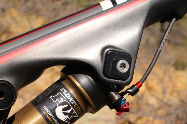 Rocky Mountain Instinct 999 MSL: Close-up of a mountain bike suspension system, highlighting the shock reservoir with branded markings. The image features a metallic frame with a sleek design, showcasing the adjustment knobs and cable connections. The background is blurred, emphasizing the detailed components of the bike.