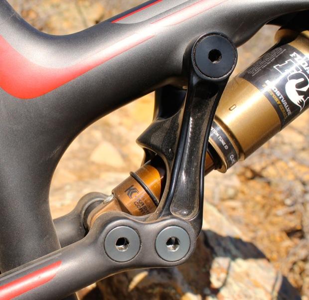 Rocky Mountain Instinct 999 MSL: Close-up view of a mountain bike suspension system, highlighting the linkage and shock absorber. The frame features a sleek design with a black and red color scheme, and the shock absorber is partially visible, showing its gold and black components against a rocky background.