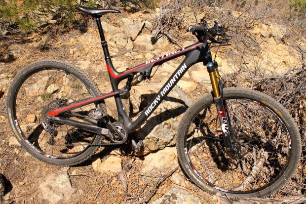 Rocky Mountain Instinct 999 MSL: A black and red Rocky Mountain mountain bike positioned on rocky terrain with sparse vegetation in the background. The bike features a suspension fork and knobby tires, indicating it's designed for off-road use.