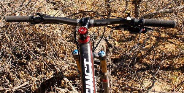 Rocky Mountain Instinct 999 MSL: Close-up view of mountain bike handlebars, showcasing the grips, brake lever, and suspension fork, set against a background of dry, brown vegetation. The bike features a distinctive brand logo prominently displayed on the frame.