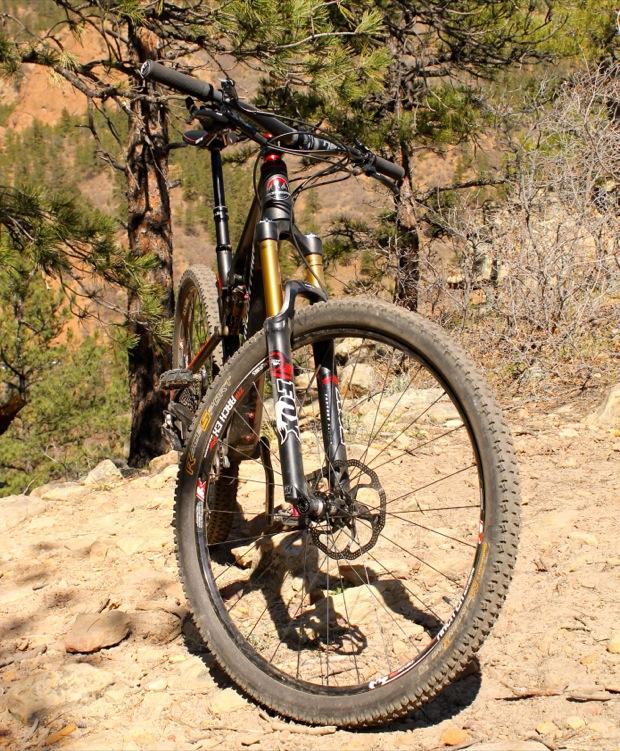 Rocky Mountain Instinct 999 MSL: Alt text: A mountain bike positioned on rocky terrain, surrounded by pine trees. The bike features a black and gold frame, thick tires, and front suspension forks. In the background, a hilly landscape is visible, indicating an outdoor setting.