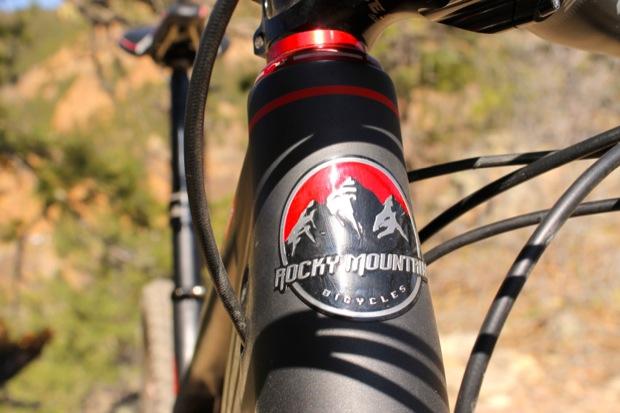 Rocky Mountain Instinct 999 MSL: Close-up view of a Rocky Mountain Bicycles logo on a mountain bike frame, showcasing the distinctive design with mountains and a red background. The image captures part of the handlebars and the natural outdoor setting with blurred greenery in the background.