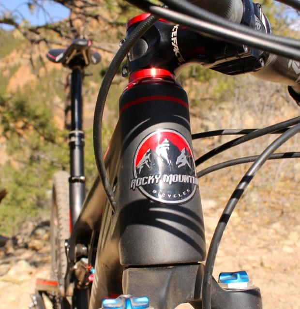 Rocky Mountain Instinct 999 MSL: Close-up view of a Rocky Mountain Bicycles logo on the frame of a mountain bike, with bike handlebars and parts visible in the background. The setting features natural outdoor elements, such as trees and rocky terrain.