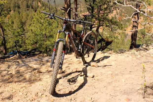 Rocky Mountain Instinct 999 MSL: A mountain bike is positioned on a dirt trail surrounded by pine trees, with a rocky terrain and mountainous landscape in the background. The bike features thick tires and a sturdy frame, suggesting it's designed for off-road cycling. The sunlight casts shadows on the ground, highlighting the bike's details.