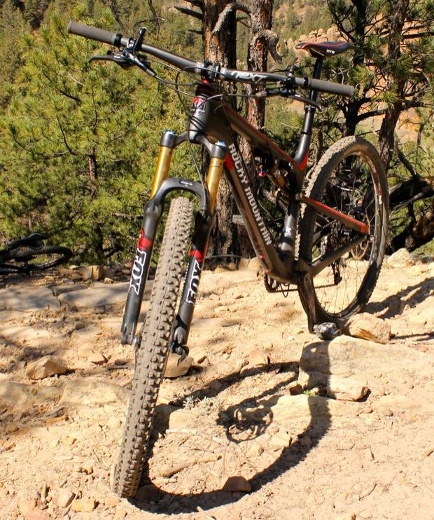 Rocky Mountain Instinct 999 MSL: A mountain bike with a black and orange frame is positioned on a rocky dirt trail surrounded by tall green trees and natural terrain. The bike features suspension forks and knobby tires, indicating it is designed for off-road riding. The sunlight highlights the bike's details and the rugged environment.