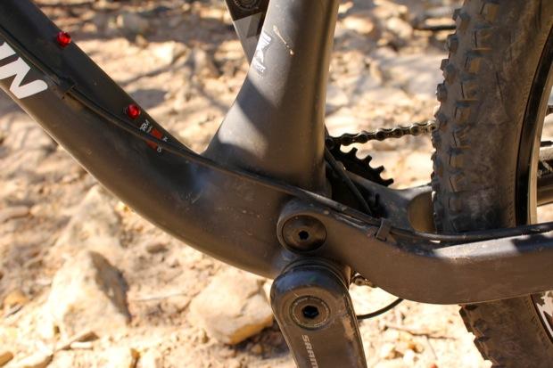 Rocky Mountain Instinct 999 MSL: Close-up view of a bicycle's bottom bracket area, showcasing the frame construction and components. The image features a black frame with visible branding, a chain, and prominent tire tread in the background, all set against a rocky surface.