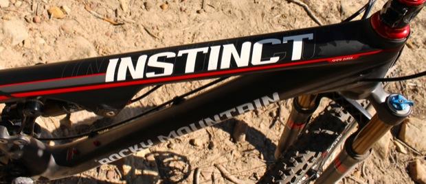 Rocky Mountain Instinct 999 MSL: A close-up view of the top tube of a Rocky Mountain Instinct mountain bike, featuring a sleek black frame with bold white and red lettering. The background shows a natural, dirt terrain, typical for outdoor biking environments.