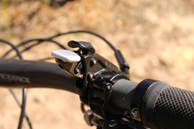 Rocky Mountain Instinct 999 MSL: Close-up view of a bicycle's handlebar with a focus on the gear shifter and grip. The metal shifter lever is positioned above a textured rubber grip, surrounded by bicycle cables. The background is softly blurred, suggesting an outdoor environment.