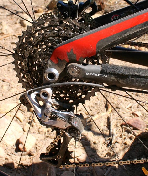 Rocky Mountain Instinct 999 MSL: Close-up view of a bicycle's rear gear system, featuring a black and red frame, a cassette of gears, and a SRAM derailleur. The background displays a natural, rocky terrain.