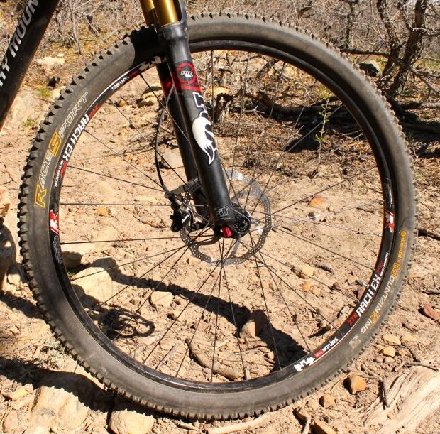 Rocky Mountain Instinct 999 MSL: Close-up of a bicycle front wheel with a black and gold suspension fork. The tire is rugged, suitable for off-road terrain, and features a disc brake. The background shows a rocky, dirt path with sparse vegetation, indicating a natural outdoor setting.