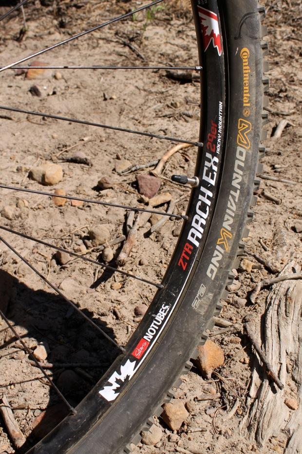 Rocky Mountain Instinct 999 MSL: Close-up of a mountain bike wheel resting on a dirt surface, showing a ZTR ARCH EX rim, a Continental mountain tire, and note of "No Tubes." Surrounding terrain includes small rocks and dry earth.