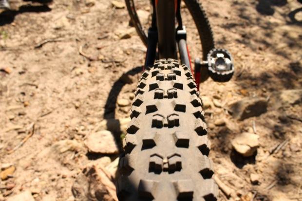 Rocky Mountain Instinct 999 MSL: Close-up view of a mountain bike tire with a distinctive knobby tread pattern, resting on a rocky dirt surface. The image captures the tire from a low angle, emphasizing its rugged design and the surrounding terrain.