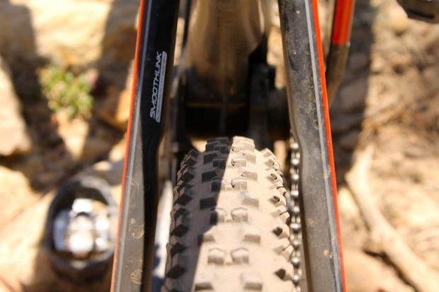 Rocky Mountain Instinct 999 MSL: Close-up view of a mountain bike rear tire and frame, highlighting the tread pattern and the "Smooth Link" branding on the bike's components. The image captures a rugged, outdoor setting with dirt and rocks in the background.