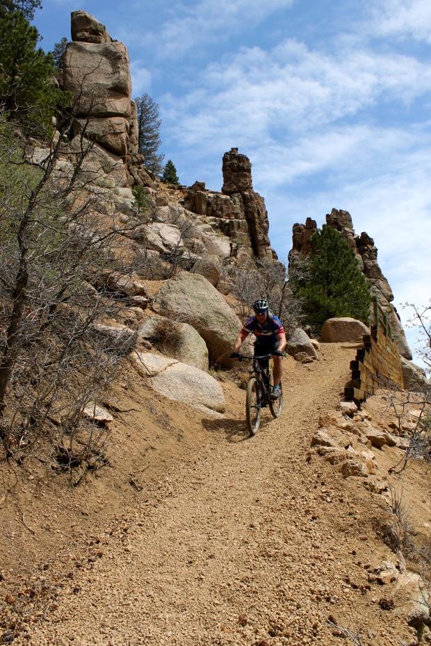 Rocky Mountain Instinct 999 MSL: A mountain biker navigating a rocky trail surrounded by tall rock formations and sparse vegetation under a partially cloudy sky.