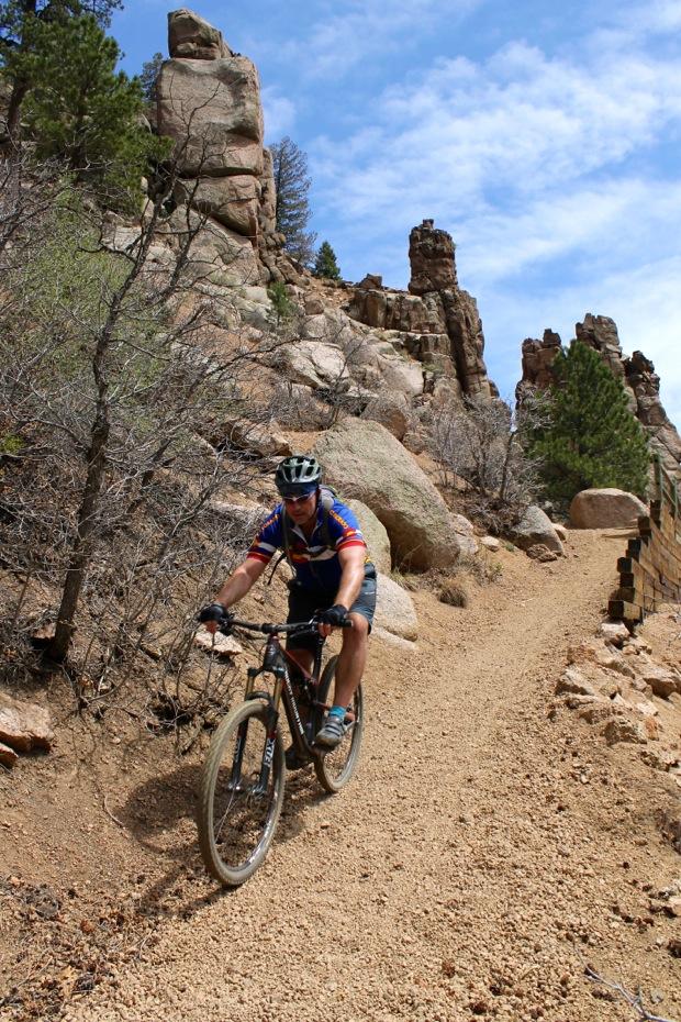 Rocky Mountain Instinct 999 MSL: A mountain biker navigating a rugged, dirt trail lined with large rocks and sparse vegetation, under a partly cloudy sky.