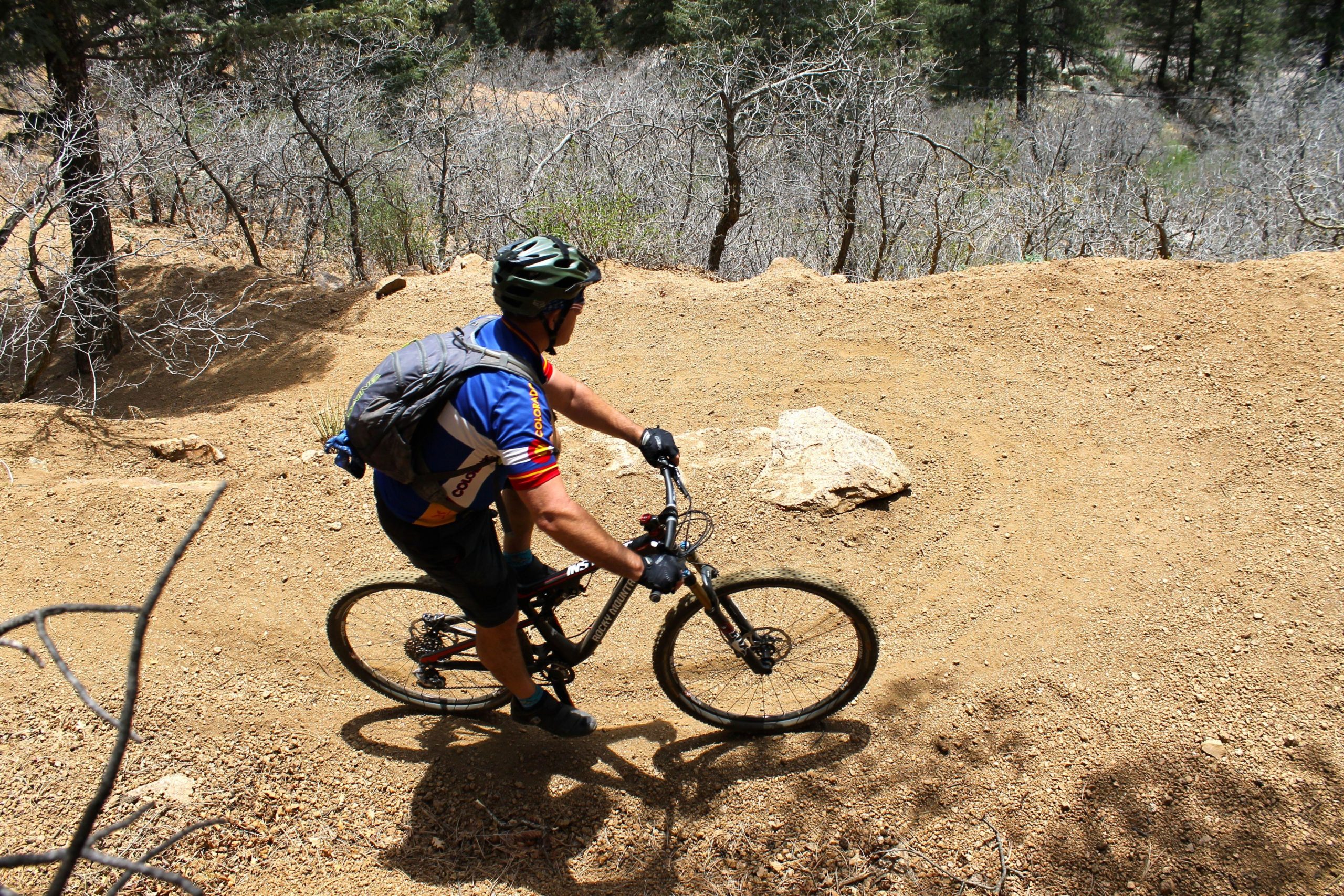 A cyclist navigating a dirt trail in a wooded area, wearing a helmet and a colorful jersey. The path is surrounded by dry vegetation and rocky terrain. The cyclist is leaning slightly to one side, indicating movement along the winding trail. Columbine mountain bike trail.