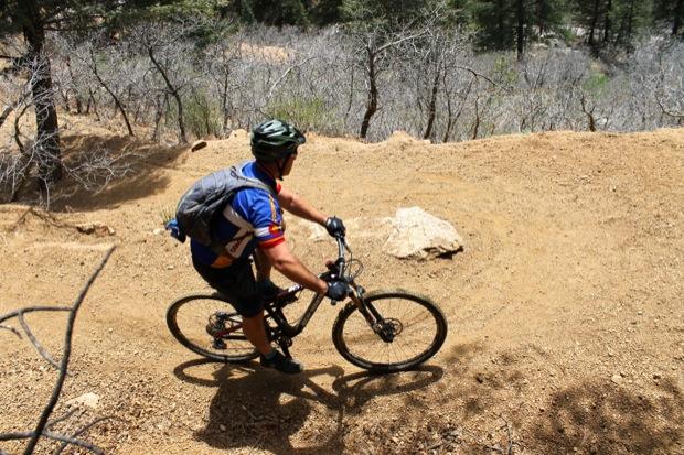 Rocky Mountain Instinct 999 MSL: A mountain biker navigating a steep, dirt trail surrounded by sparse trees and underbrush. The rider is wearing a blue jersey, a helmet, and a backpack, focused on maneuvering around a rocky outcrop. The scene captures the thrill of outdoor biking in a natural setting.