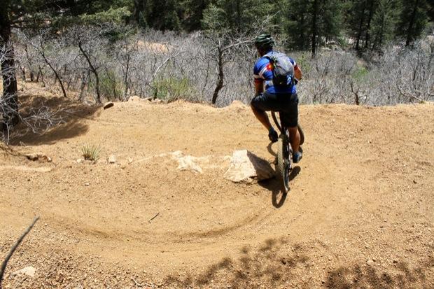 Rocky Mountain Instinct 999 MSL: A mountain biker wearing a blue jersey and helmet rides along a dirt trail that curves around a rock. The surrounding area consists of sparse trees and dry vegetation, indicating a rugged outdoor setting.