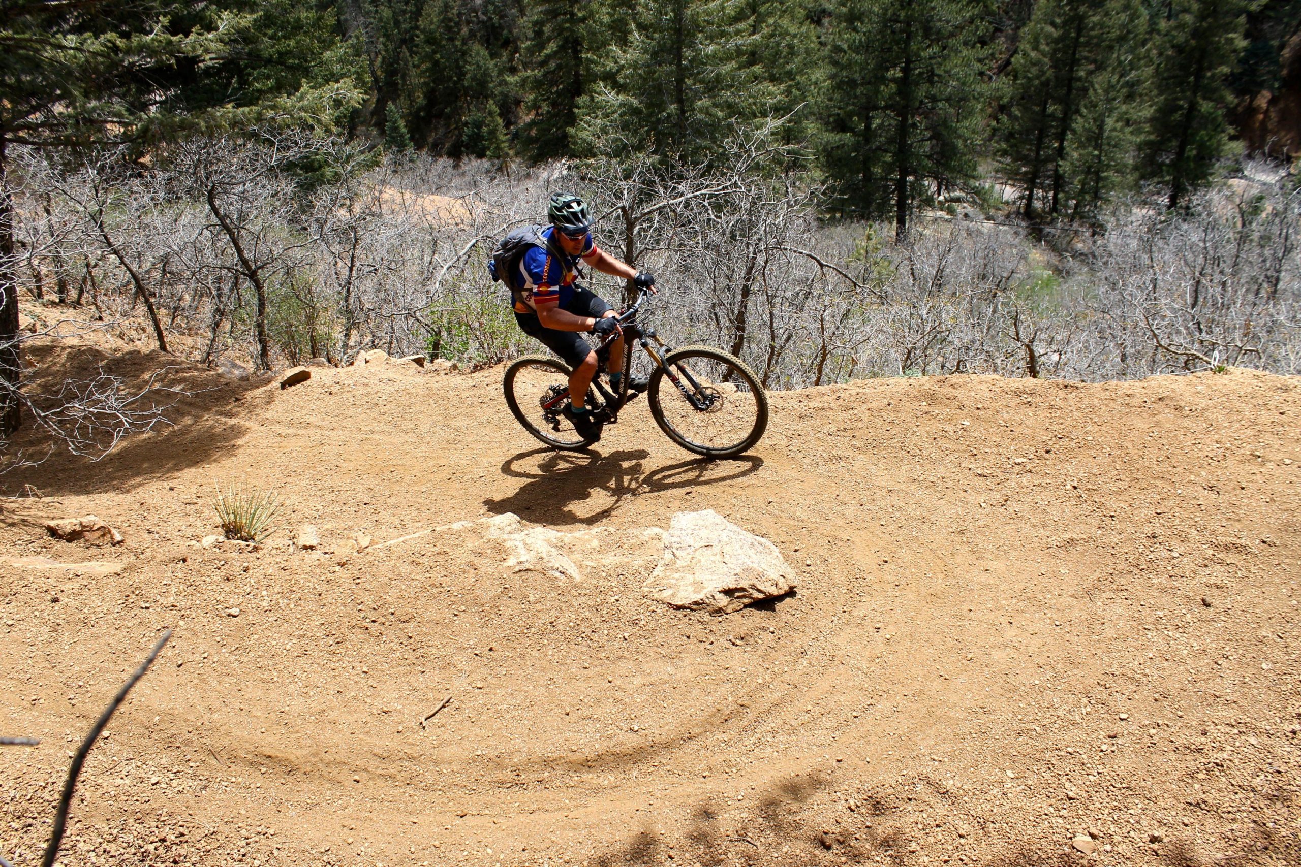 A mountain biker navigating a dirt trail while leaning into a turn, surrounded by trees and sparse vegetation. The terrain is rocky and dry, showcasing a recreational outdoor setting. Columbine mountain bike trail.