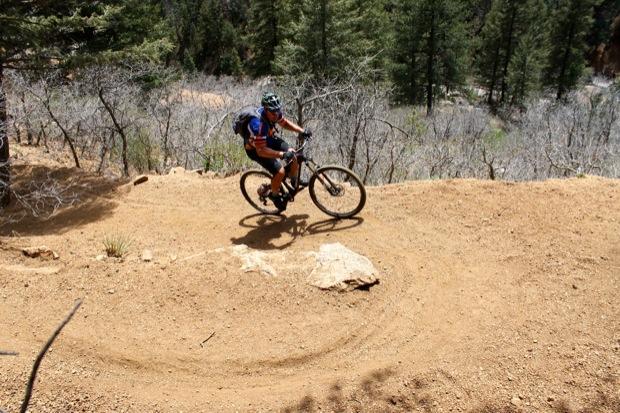 Rocky Mountain Instinct 999 MSL: A mountain biker navigates a dirt trail, riding around a curved path on a sunny day. The surrounding landscape features sparse trees and rocky terrain, highlighting the rugged outdoor environment.