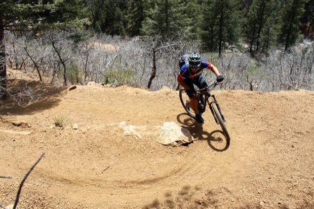 Rocky Mountain Instinct 999 MSL: A mountain biker navigating a curved dirt trail on a rocky hillside, surrounded by trees and sparse vegetation. The cyclist is wearing a helmet and a brightly colored jersey, with a backpack visible on their back.